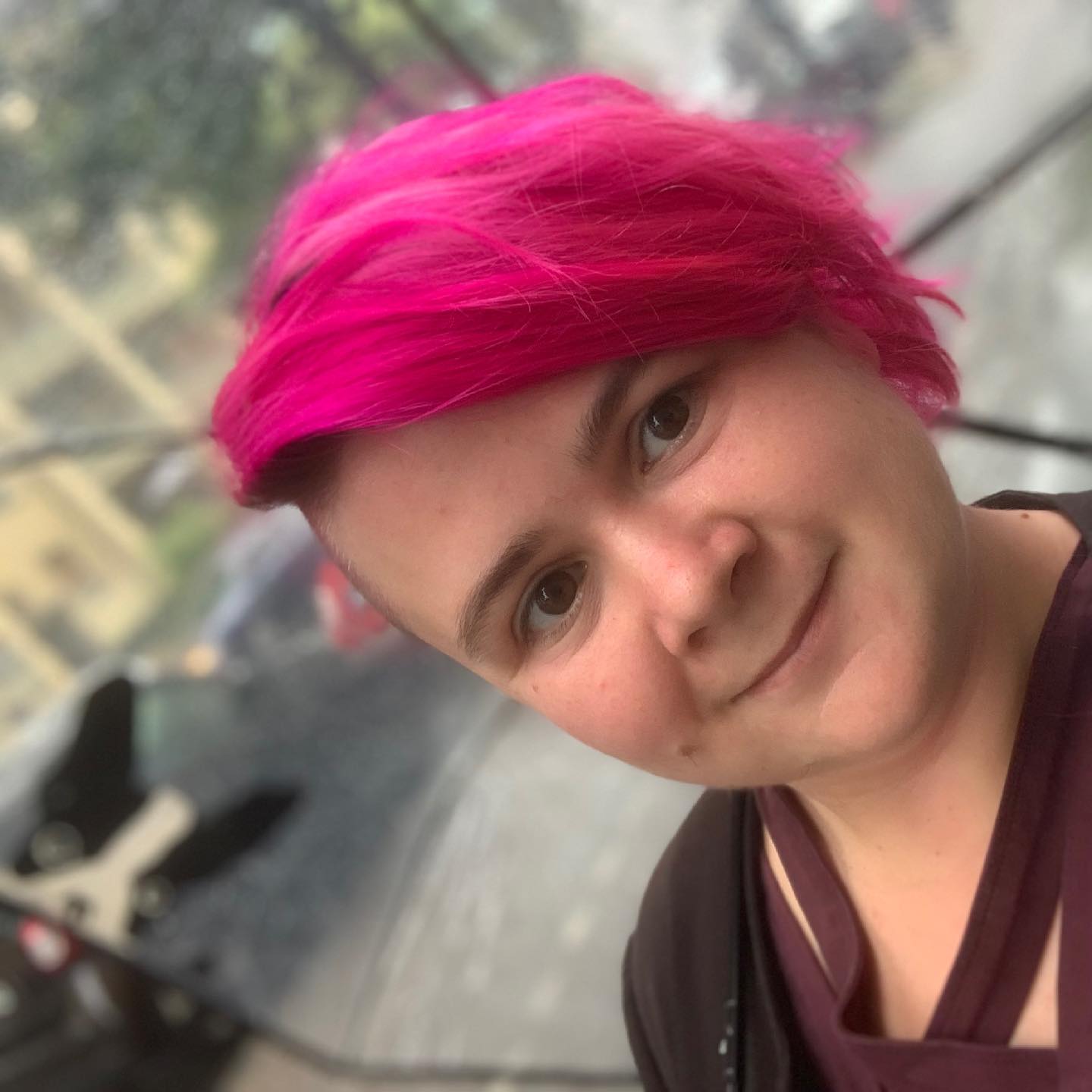 Portrait picture of a cis female with pink hair, pixie cut, smiling at the camera.
