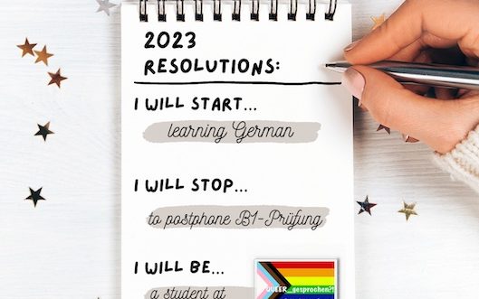 a little notebook on a white wooden table and glitter stars. Text on the notebook says: 2023 resolutions: I will start learning German. I will stop to postphone B1-Prüfung. I will be a student at QUEER_gesprochen?!